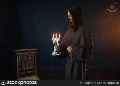 Medieval monk in robe holds a candlestick in hands, black background, secret ritual. Mysterious friar with candles. Mystery and spirituality. Medieval monk in robe holds a candlestick in hands