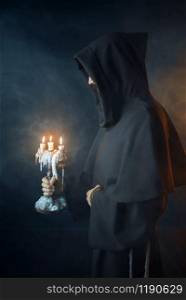 Medieval monk in robe holds a candlestick in hands, black background, secret ritual. Mysterious friar with candles. Mystery and spirituality. Medieval monk in robe holds a candlestick in hands