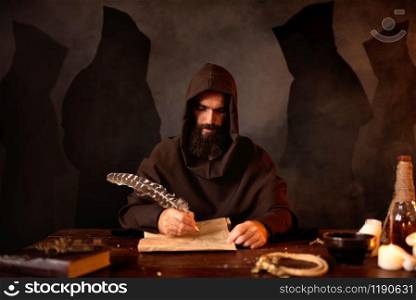 Medieval monk in robe makes notes in the secret scripture with a goose feather, black background, secret ritual. Mysterious friar in dark cape. Mystery and spirituality. Medieval monk makes notes with a goose feather