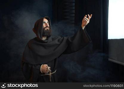 Medieval monk praying against a window with bright light, religion. Mysterious friar in dark cape, Mystery and spirituality