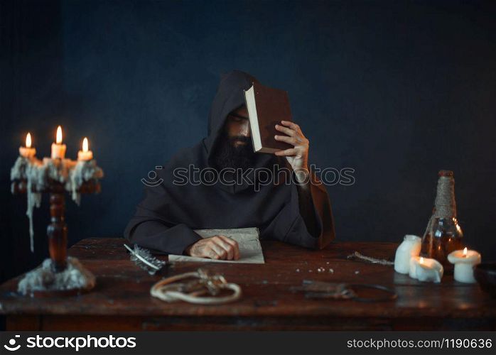 Medieval monk sitting at the table and reads secret scripture. Mysterious friar in dark cape. Mystery and spirituality