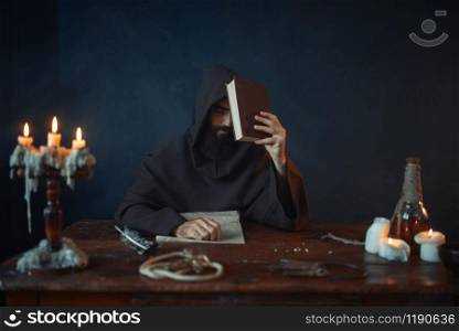 Medieval monk sitting at the table and reads secret scripture. Mysterious friar in dark cape. Mystery and spirituality
