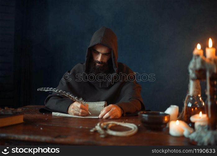 Medieval monk sitting at the table and writes with a goose feather, top view, secret scripture. Mysterious friar in dark cape. Mystery and spirituality. Medieval monk sitting at table and write, top view