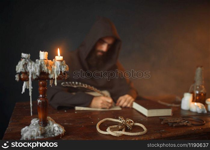 Medieval monk sitting at the table and writes with a goose feather, top view, secret scripture. Mysterious friar in dark cape. Mystery and spirituality. Medieval monk sitting at table and write, top view