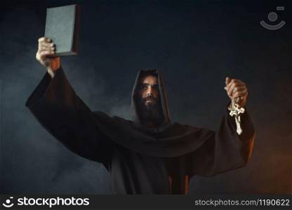 Medieval monk stands in fire with book in hands, black background, secret ritual. Mysterious friar in dark cape. Mystery and spirituality. Medieval monk stands in fire with book in hands