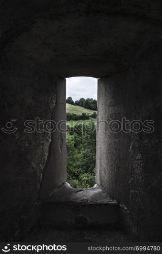 Medieval stone window and green landscape with forest and meadow viewed through it. Dramatic light. Stone window frame. Old German architecture.