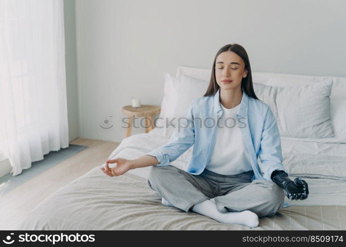 Meditation and concentration. Morning routine of disabled woman. Girl is sitting in lotus pose in her bedroom. Attractive european woman has artificial cyber limb. Stress relief at home.. Meditation and concentration. Morning routine of disabled woman with artificial cyber limb.