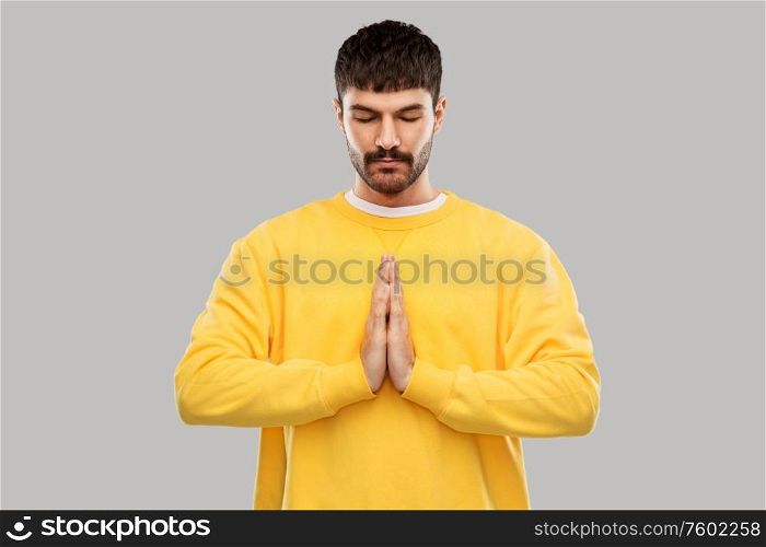 meditation, faith and mindfulness concept - young man in yellow sweatshirt meditating or praying over grey background. man in yellow sweatshirt meditating or praying