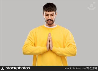 meditation, faith and mindfulness concept - young man in yellow sweatshirt meditating or praying over grey background. man in yellow sweatshirt meditating or praying