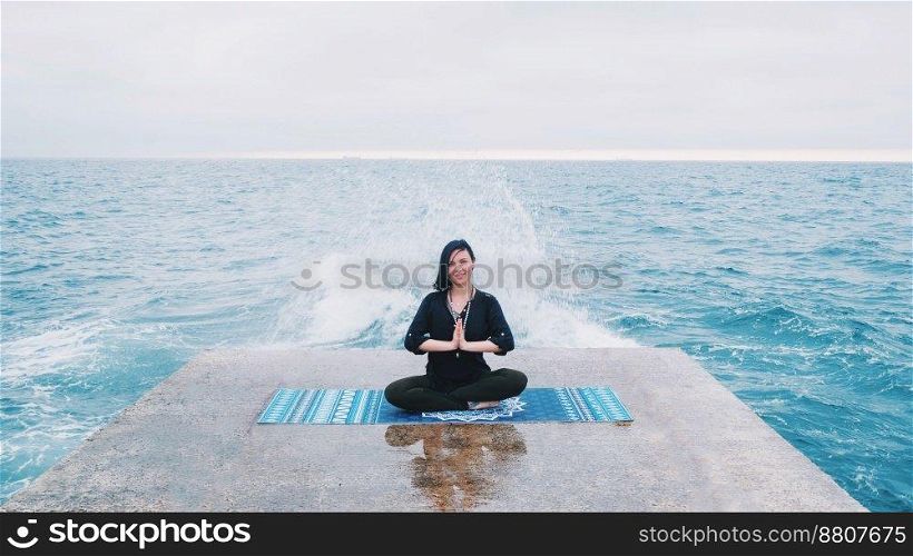 Meditation. Yoga young calm brunette woman by the waves sea on pier relaxing in serene zen. lotus yoga pose on a beach at cloudy weather. Beautiful colorful background.