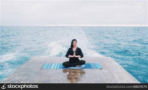 Meditation. Yoga young calm brunette woman by the waves sea on pier relaxing in serene zen. lotus yoga pose on a beach at cloudy weather. Beautiful colorful background.