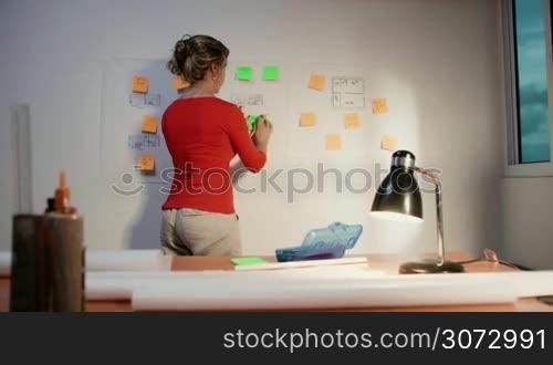 Medium shot of blond woman working as architect, sticking an adhesive note to a blueprint. She sits back and contemplates the missing tasks to complete the housing project. Dolly shot