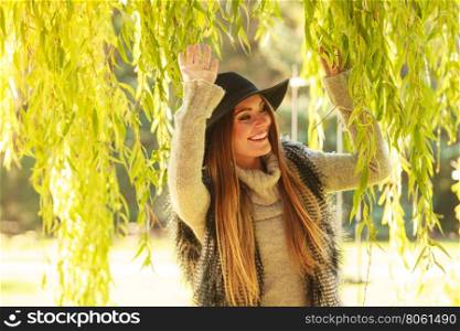 Meeting time. Trendy young woman waiting for someone in park waving her hand around green leaves of willow tree.