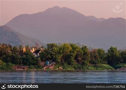 Mekhong or Mae Khong river view and Old aged Buddhist temple with lush green forest and mountains in Luang Prabang, Laos under evening sky