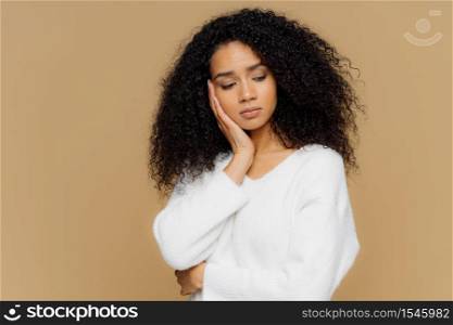 Melancholic sad lonely woman focused down with thoughtful expression, keeps hand on cheek, has calm expression, wears white sweater, has curly hair, isolated on brown studio wall. Depression