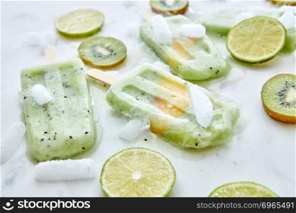 Melting green berry ice cream lolly with a piece of peach on a white background with juicy pieces of kiwi, lime and ice. Cold dessert. Flat lay. Frozen fruit smoothies with a slice of mango presented on a gray background with pieces of ice, kiwi and lime. Healthy summer dessert. Flat lay