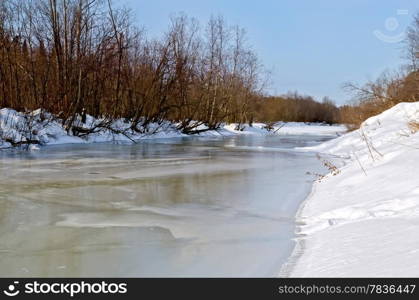 Melting ice on the river, snow, bushes and trees on the shore against the blue sky