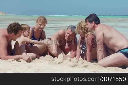 Members of a big family are resting on the beach. They are building a sand castle and talking to each other.
