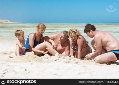 Members of a big family are sitting by the sea and building sand castle.