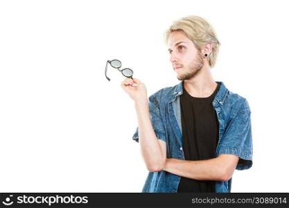 Men fashion, clothing, accessories concept. Artistic hipster guy holding glasses wearing jeans outfit looking into space and thinking, studio shot isolated.. Artistic hipster guy holding glasses