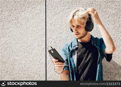 Men fashion, technology, urban style clothing concept. Hipster guy standing on city street wearing jeans outfit and sunglasses listening to music and holding phone. Hipster man listening music through headphones