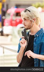 Men fashion, technology, urban style clothing concept. Hipster guy standing on city street wearing jeans outfit listening to music through earphones. Hipster man standing on city street listening music