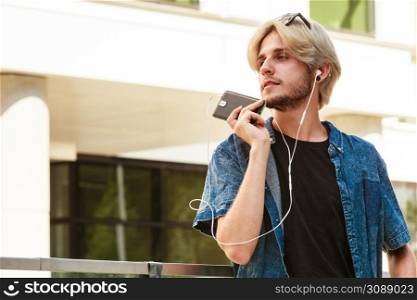 Men fashion, technology, urban style clothing concept. Hipster guy standing on city street wearing jeans outfit listening to music through earphones, talking on phone. Hipster man standing with earphones talking on phone