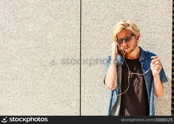 Men fashion, technology, urban style clothing concept. Hipster guy standing on city street wearing jeans outfit and eccentric sunglasses listening to music and holding phone. Hipster man listening music through earphones