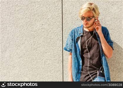 Men fashion, technology, urban style clothing concept. Hipster guy standing on city street wearing jeans outfit and eccentric sunglasses listening to music and holding phone. Hipster man listening music through earphones