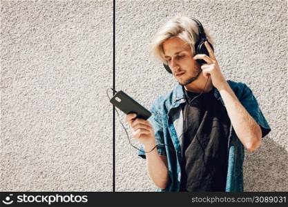 Men fashion, technology, urban style clothing concept. Hipster guy standing on city street wearing jeans outfit and sunglasses listening to music and holding phone. Hipster man listening music through headphones