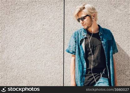 Men fashion, technology, urban style clothing concept. Hipster guy standing on city street wearing jeans outfit and eccentric sunglasses listening to music and holding phone. Hipster man listening music through earphones
