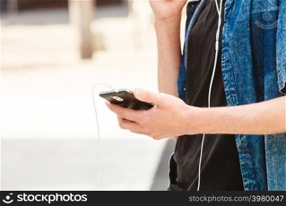Men fashion, technology, urban style clothing concept. Hipster guy standing on city street wearing jeans outfit holding smartphone with earphones, no face. Man in jeans shirt holding moblie phone