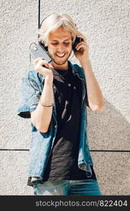 Men fashion, technology, urban style clothing concept. Hipster guy standing on city street wearing jeans outfit and sunglasses listening to music and holding phone. Hipster smiling man listening music through headphones