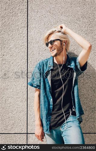 Men fashion, technology, urban style clothing concept. Hipster smiling guy standing on city street wearing jeans outfit listening to music through earphones. Hipster man standing on city street listening music