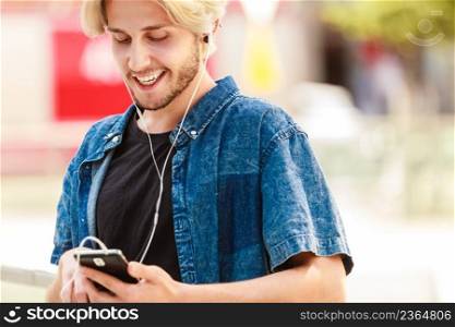 Men fashion, technology, urban style clothing concept. Hipster smiling guy standing on city street wearing jeans outfit listening to music through earphones. Hipster man standing on city street listening music
