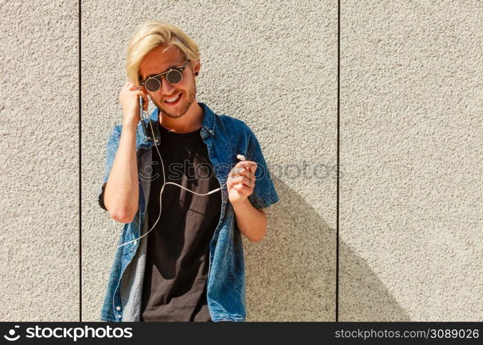 Men fashion, technology, urban style clothing concept. Hipster smiling guy standing on city street wearing jeans outfit and sunglasses listening to music through earphones. Hipster man smiling listening music through earphones