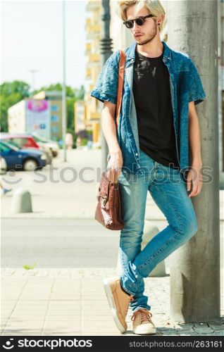 Men fashion, urban style clothing concept. Hipster guy walking through city wearing jeans outfit, male handbag and sunglasses on sunny day. Hipster man standing on city street, urban fashion