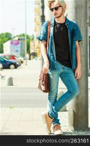Men fashion, urban style clothing concept. Hipster guy walking through city wearing jeans outfit, male handbag and sunglasses on sunny day. Hipster man standing on city street, urban fashion
