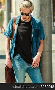 Men fashion, urban style clothing concept. Hipster guy walking through city wearing jeans outfit, male handbag and sunglasses on sunny day. Hipster man walking through city, urban style