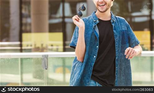 Men fashion, urban style clothing concept. Hipster smiling guy standing on city street wearing jeans outfit, male handbag, holding sunglasses on sunny day. Hipster man standing on city street, urban fashion