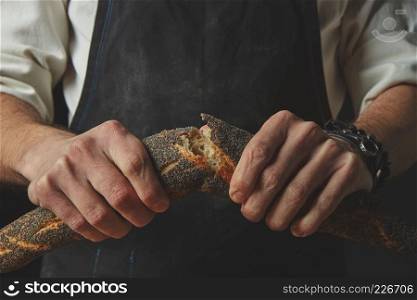 Men Hand breaking and separating a freshly baked baguette with poppy seeds on a dark background. Concept of bread.. hands men break the baguette