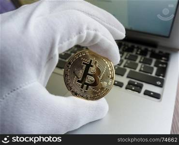Men in white glove holding golden bitcoin against laptop. Focus on the coin.
