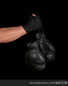 men's arm wrapped in a black elastic sports bandage holds pair old vintage leather boxing gloves, black background, copy space