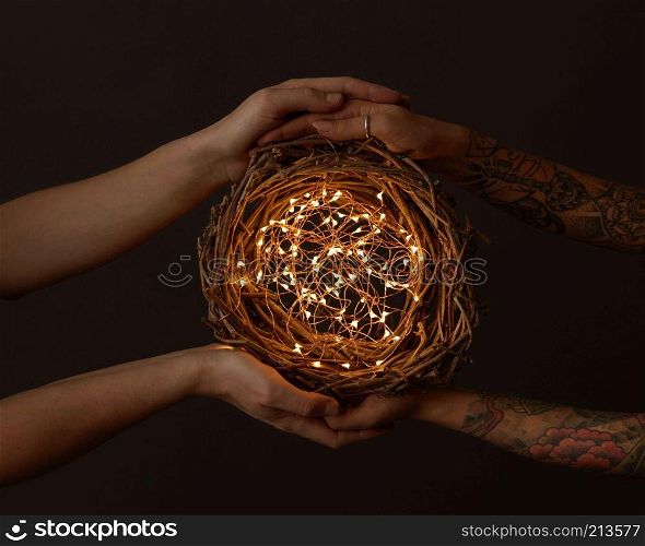 Men&rsquo;s hands and women&rsquo;s hands with a tattoo holding a wreath of branches with yellow garlands around a dark background.. A wreath of branches with bright garlands are held by male and female hands with a tattoo on a black background. Celebration