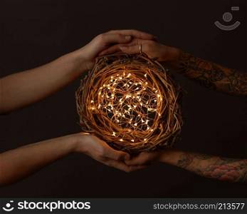 Men&rsquo;s hands and women&rsquo;s hands with a tattoo holding a wreath of branches with yellow garlands around a dark background.. A wreath of branches with bright garlands are held by male and female hands with a tattoo on a black background. Celebration