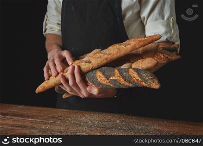 Men&rsquo;s hands hold freshly baked baguettes against the black background of a wooden table. Men&rsquo;s hands hold baguettes