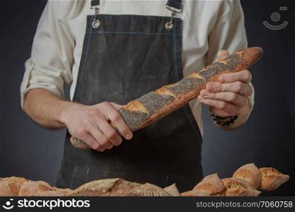 Men&rsquo;s hands holding a fresh baguette with poppy seeds on a dark background clous up. Men&rsquo;s hands hold a baguette