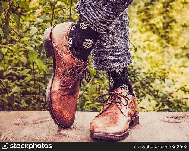 Men's legs in stylish shoes, black socks with patterns in the form of US dollars on a wooden terrace against the background of green trees. Beauty, fashion, elegance. Men's legs in stylish shoes, black socks with patterns
