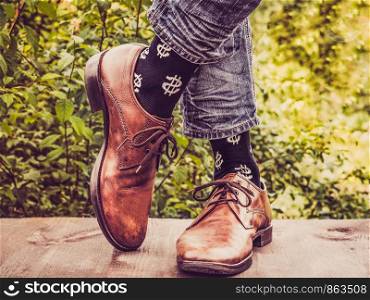 Men's legs in stylish shoes, black socks with patterns in the form of US dollars on a wooden terrace against the background of green trees. Beauty, fashion, elegance. Men's legs in stylish shoes, black socks with patterns