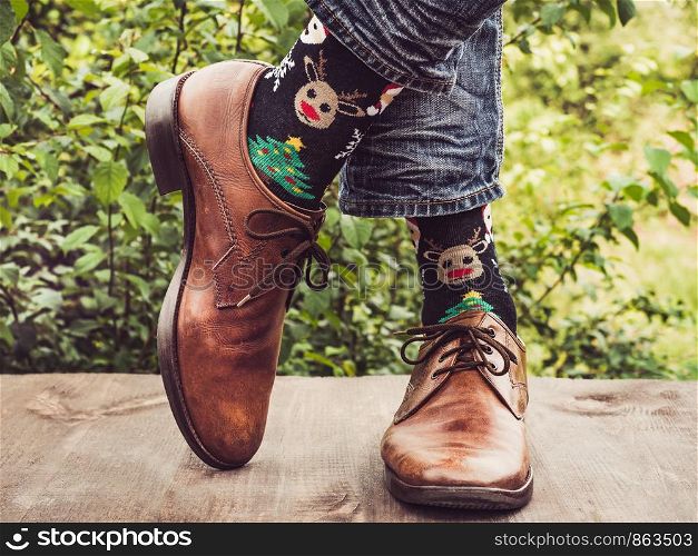 Men's legs in stylish shoes, bright, variegated socks with Christmas and New Year's patterns on the wooden terrace on the background of green trees. Beauty, fashion, elegance. Men's legs in stylish shoes, bright, variegated socks